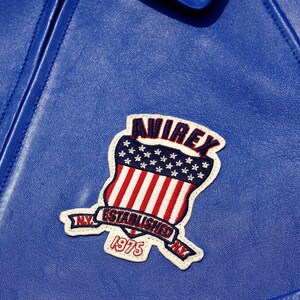 K&ouml;nnte beinhalten: Nahaufnahme einer blauen Lederjacke mit einem aufgen&auml;hten Aufn&auml;her. Der Aufn&auml;her zeigt ein Schilddesign mit roten und wei&szlig;en Streifen, blauen Sternen und dem Wort "AVIREX" &uuml;ber dem Schild. Der Aufn&auml;her enth&auml;lt auch den Text "ESTABLISHED 1975" und "NY".