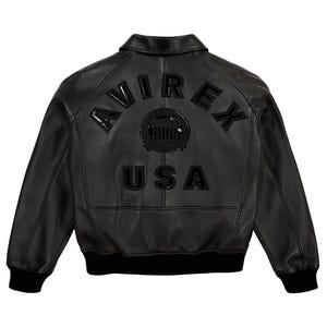 Puede incluir: Chaqueta bomber de cuero negro con la palabra "AVIREX" arqueada en la parte superior de la espalda, encima de un emblema circular y la palabra "USA". La chaqueta tiene cuello, pu&ntilde;os y dobladillo de canal&eacute;.