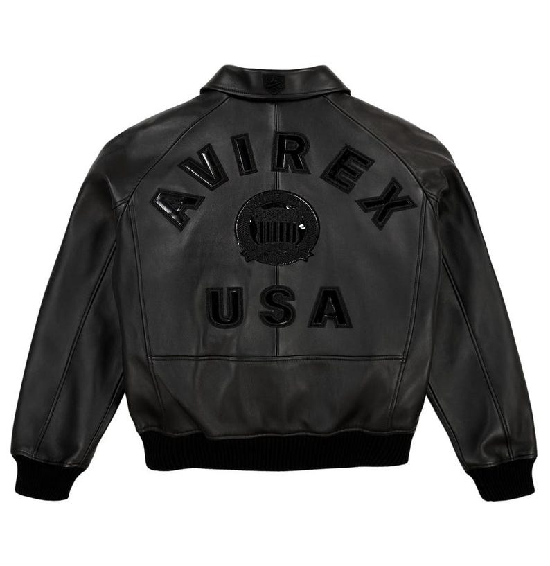 Puede incluir: Chaqueta bomber de cuero negro con la palabra "AVIREX" arqueada en la parte superior de la espalda, encima de un emblema circular y la palabra "USA". La chaqueta tiene cuello, pu&ntilde;os y dobladillo de canal&eacute;.