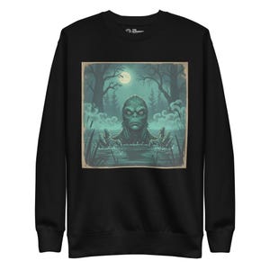 May include: Black sweatshirt with a vintage illustration of a swamp creature rising from water under a full moon. The artwork is framed, set against a backdrop of a dark forest and misty water.