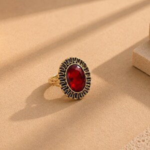 May include: A gold-toned ring featuring a large, oval-shaped, faceted deep red gemstone at its centre. The stone is surrounded by a decorative, scalloped metal border with a textured, wavy pattern that frames the central red jewel.