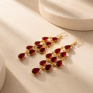Peut inclure: Une paire de boucles d'oreilles pendantes dorées composées de multiples pierres rouge foncé en forme de goutte, disposées en cascade sur plusieurs niveaux. Chaque boucle est suspendue à un crochet doré simple au fini métallique poli.