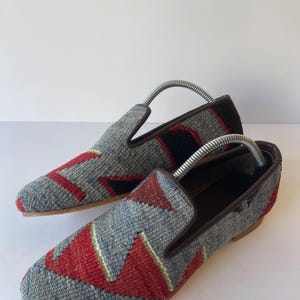 Pode incluir: Um par de mocassins estampados com um design geom&eacute;trico cinza, vermelho e preto. Os sapatos t&ecirc;m acabamentos e solas em couro castanho. S&atilde;o inseridas formas para sapatos. Cerca de 25 cm de comprimento.