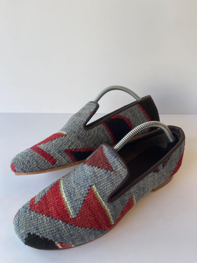 Pode incluir: Um par de mocassins estampados com um design geom&eacute;trico cinza, vermelho e preto. Os sapatos t&ecirc;m acabamentos e solas em couro castanho. S&atilde;o inseridas formas para sapatos. Cerca de 25 cm de comprimento.