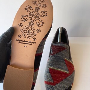 Pode incluir: Um par de sapatos feitos &agrave; m&atilde;o com cabedal de kilim e solas de couro genu&iacute;no. Os sapatos apresentam um padr&atilde;o geom&eacute;trico cinza, vermelho e preto na parte superior. A sola &eacute; bege com um design decorativo e o texto "Vintage handmade kilim upper Genuine leather sole 42".