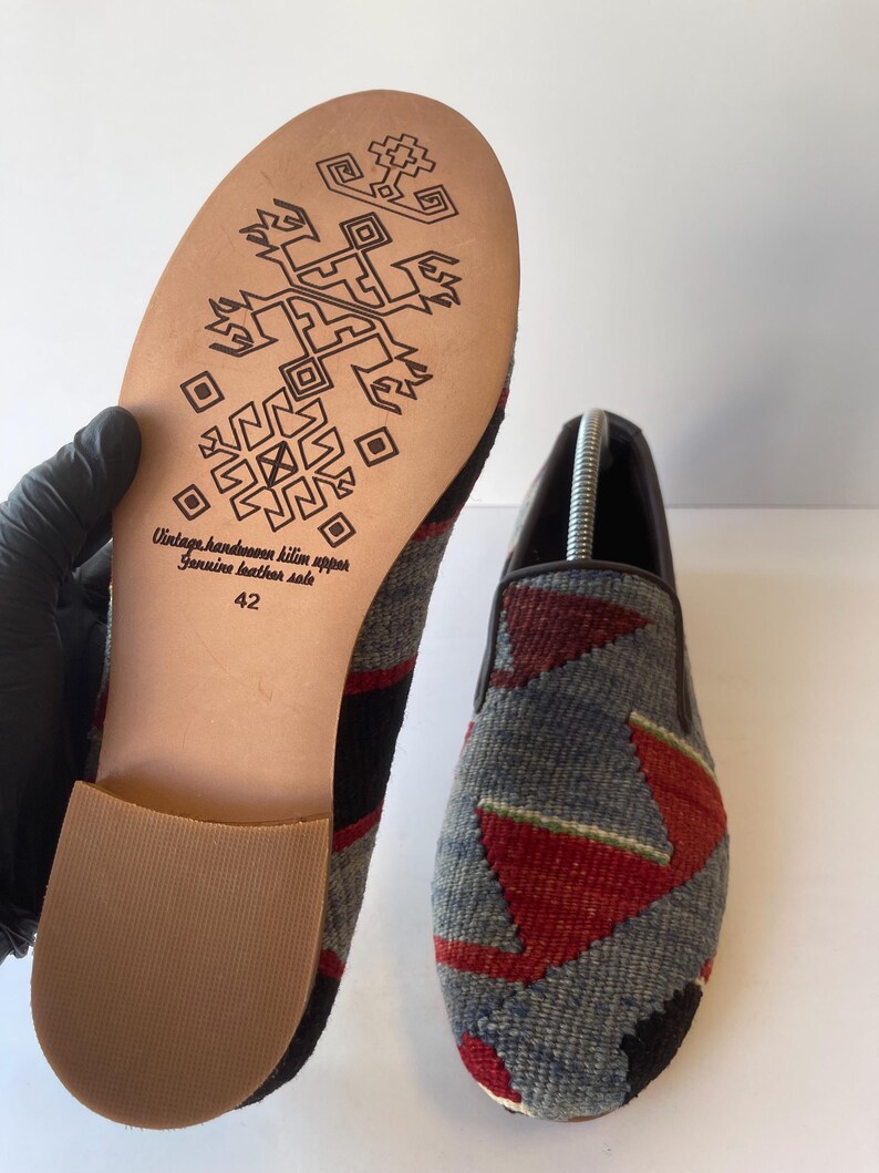 Pode incluir: Um par de sapatos feitos &agrave; m&atilde;o com cabedal de kilim e solas de couro genu&iacute;no. Os sapatos apresentam um padr&atilde;o geom&eacute;trico cinza, vermelho e preto na parte superior. A sola &eacute; bege com um design decorativo e o texto "Vintage handmade kilim upper Genuine leather sole 42".