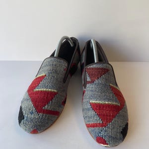 Pode incluir: Um par de sapatos cinzentos sem atacadores com um design t&ecirc;xtil tecido. Os sapatos apresentam padr&otilde;es geom&eacute;tricos vermelhos e pretos. As solas s&atilde;o castanhas e o interior &eacute; escuro. S&atilde;o inseridas formas de sapatos.
