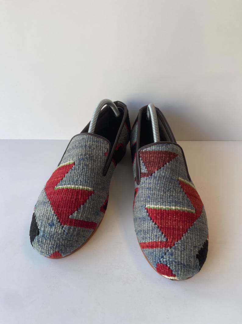 Pode incluir: Um par de sapatos cinzentos sem atacadores com um design t&ecirc;xtil tecido. Os sapatos apresentam padr&otilde;es geom&eacute;tricos vermelhos e pretos. As solas s&atilde;o castanhas e o interior &eacute; escuro. S&atilde;o inseridas formas de sapatos.