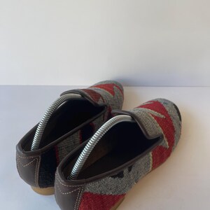 Pode incluir: Par de mocassins tecidos com um padr&atilde;o geom&eacute;trico em vermelho, cinza e preto. Os sapatos t&ecirc;m acabamentos e solas em couro marrom. As formas s&atilde;o inseridas nos sapatos para manter sua forma. Os sapatos s&atilde;o exibidos em uma superf&iacute;cie branca.