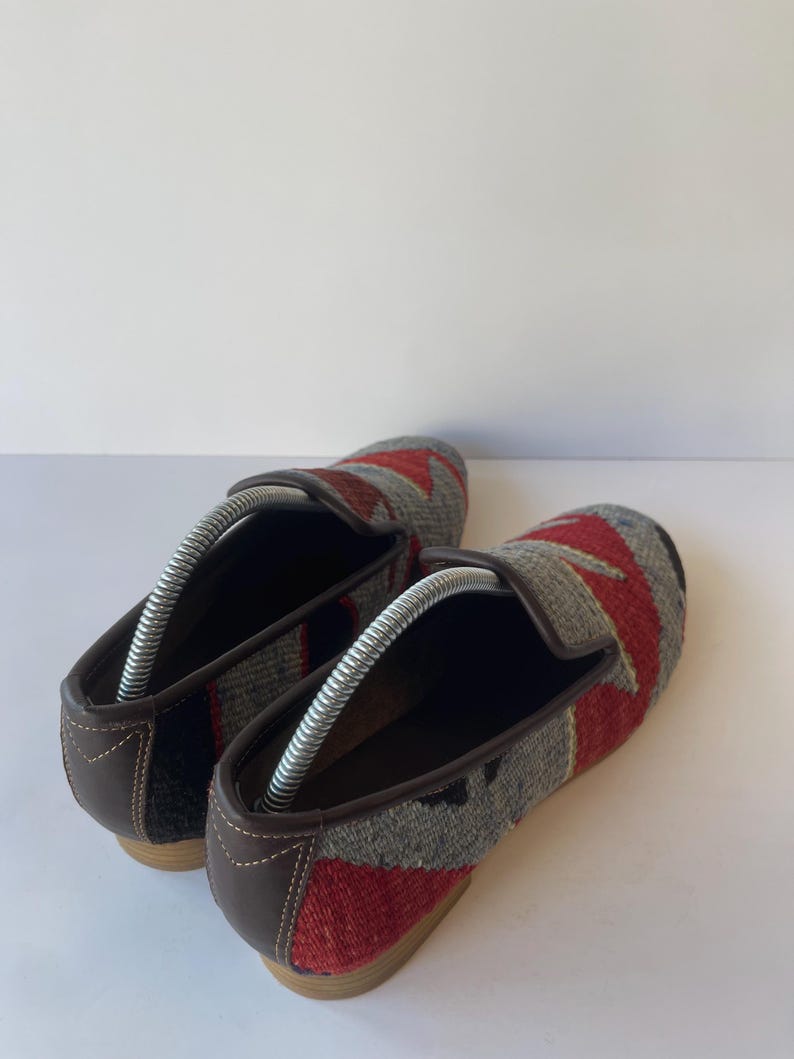 Pode incluir: Par de mocassins tecidos com um padr&atilde;o geom&eacute;trico em vermelho, cinza e preto. Os sapatos t&ecirc;m acabamentos e solas em couro marrom. As formas s&atilde;o inseridas nos sapatos para manter sua forma. Os sapatos s&atilde;o exibidos em uma superf&iacute;cie branca.