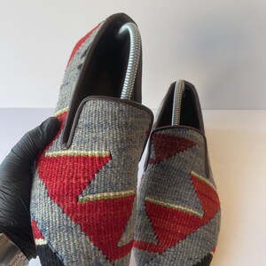 Pode incluir: Um par de sapatos cinzentos sem atacadores com um padr&atilde;o geom&eacute;trico em vermelho, preto e verde. Os sapatos t&ecirc;m uma borda castanha e s&atilde;o feitos de tecido tecido. As formas de sapato s&atilde;o inseridas no interior.