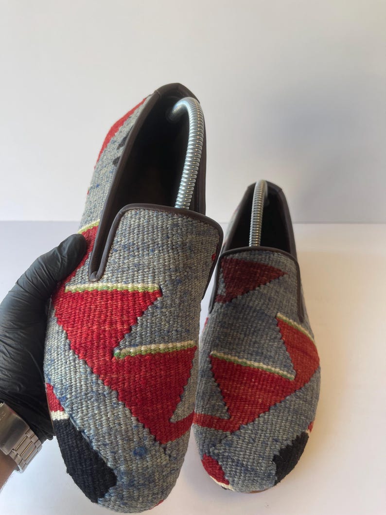 Pode incluir: Um par de sapatos cinzentos sem atacadores com um padr&atilde;o geom&eacute;trico em vermelho, preto e verde. Os sapatos t&ecirc;m uma borda castanha e s&atilde;o feitos de tecido tecido. As formas de sapato s&atilde;o inseridas no interior.