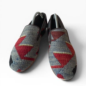 Pode incluir: Par de sapatos slip-on com cabedal em tecido. Os sapatos apresentam um fundo cinza com padr&otilde;es geom&eacute;tricos vermelhos e azuis. A guarni&ccedil;&atilde;o e as solas s&atilde;o castanhas e a biqueira &eacute; preta.