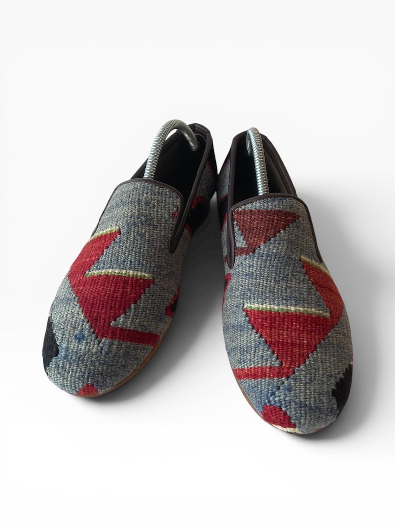 Pode incluir: Par de sapatos slip-on com cabedal em tecido. Os sapatos apresentam um fundo cinza com padr&otilde;es geom&eacute;tricos vermelhos e azuis. A guarni&ccedil;&atilde;o e as solas s&atilde;o castanhas e a biqueira &eacute; preta.