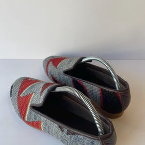 Pode incluir: Um par de sapatos slip-on cinza, vermelho e azul estampados. Os sapatos t&ecirc;m um design geom&eacute;trico na parte superior, com acabamentos e solas em couro marrom. As formas de sapato s&atilde;o inseridas.