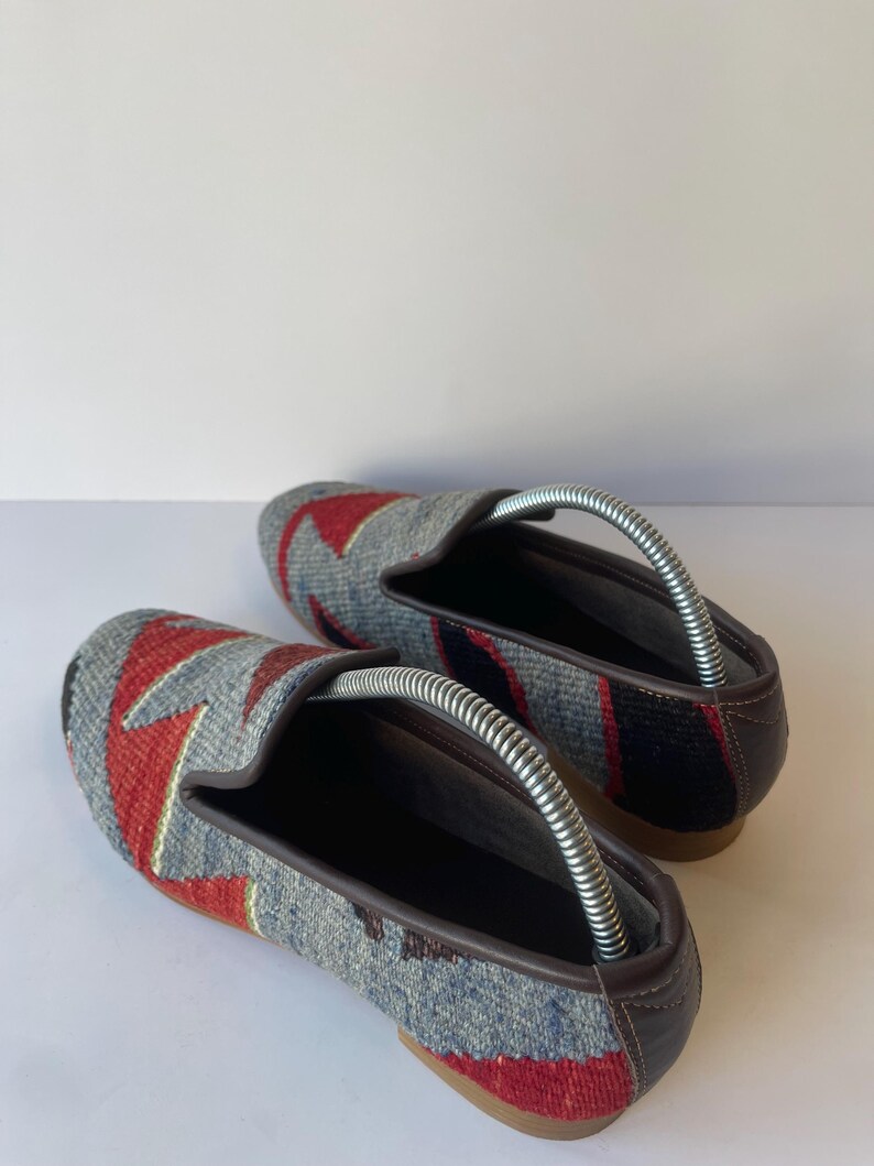 Pode incluir: Um par de sapatos slip-on cinza, vermelho e azul estampados. Os sapatos t&ecirc;m um design geom&eacute;trico na parte superior, com acabamentos e solas em couro marrom. As formas de sapato s&atilde;o inseridas.