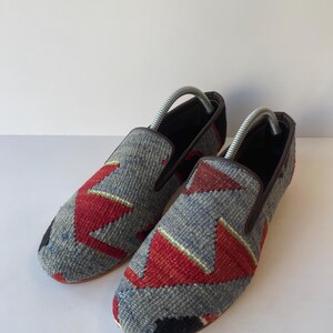 Pode incluir: Um par de sapatos slip-on com cabedal em tecido. Os sapatos apresentam um padr&atilde;o geom&eacute;trico cinza, vermelho e preto. A guarni&ccedil;&atilde;o &eacute; marrom e o interior &eacute; preto. As formas de sapato s&atilde;o inseridas.