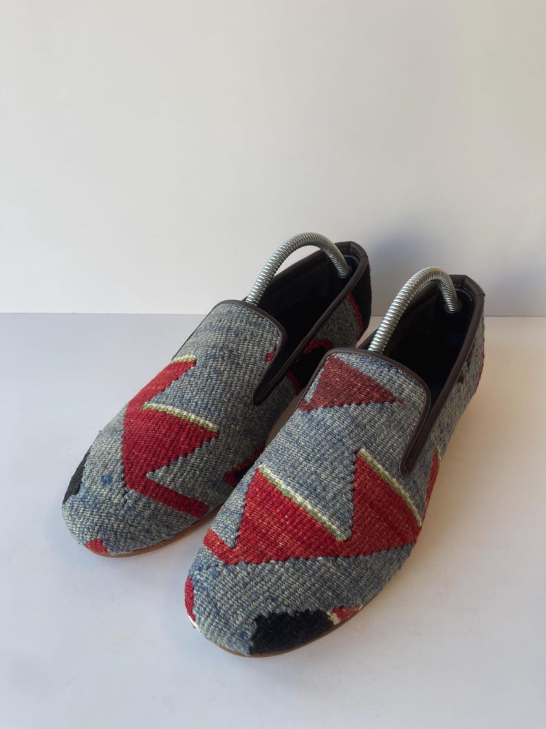 Pode incluir: Um par de sapatos slip-on com cabedal em tecido. Os sapatos apresentam um padr&atilde;o geom&eacute;trico cinza, vermelho e preto. A guarni&ccedil;&atilde;o &eacute; marrom e o interior &eacute; preto. As formas de sapato s&atilde;o inseridas.