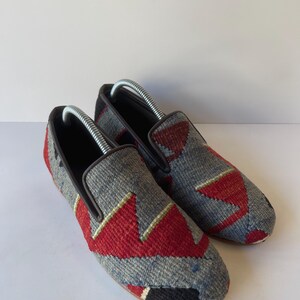 Pode incluir: Um par de sapatos slip-on cinza e vermelho estampados. Os sapatos t&ecirc;m um design geom&eacute;trico com detalhes em vermelho e preto. Eles t&ecirc;m uma borda marrom e um interior preto. As formas de sapato s&atilde;o inseridas.