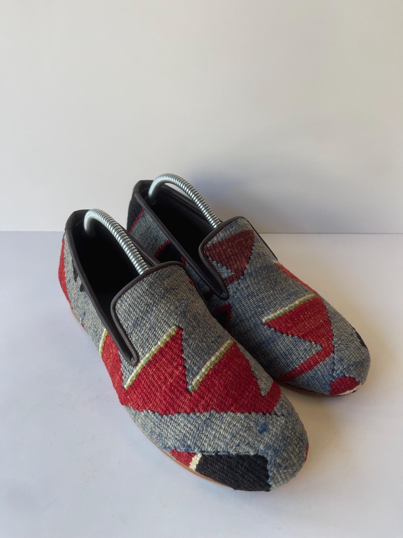 Pode incluir: Um par de sapatos slip-on cinza e vermelho estampados. Os sapatos t&ecirc;m um design geom&eacute;trico com detalhes em vermelho e preto. Eles t&ecirc;m uma borda marrom e um interior preto. As formas de sapato s&atilde;o inseridas.