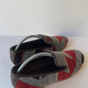 Pode incluir: Par de sapatos slip-on tecidos com um padr&atilde;o geom&eacute;trico em vermelho, cinza e preto. Os sapatos t&ecirc;m acabamentos e solas em couro marrom, e formas de metal s&atilde;o inseridas no interior. Os sapatos s&atilde;o exibidos em uma superf&iacute;cie branca.