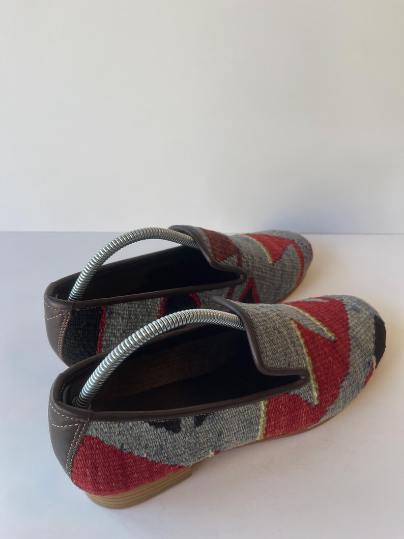 Pode incluir: Par de sapatos slip-on tecidos com um padr&atilde;o geom&eacute;trico em vermelho, cinza e preto. Os sapatos t&ecirc;m acabamentos e solas em couro marrom, e formas de metal s&atilde;o inseridas no interior. Os sapatos s&atilde;o exibidos em uma superf&iacute;cie branca.