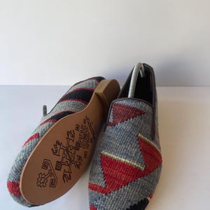 Pode incluir: Um par de sapatos slip-on tecidos com um padr&atilde;o geom&eacute;trico em cinza, vermelho e preto. As solas s&atilde;o de cor castanho claro com um design decorativo castanho escuro. Os sapatos s&atilde;o exibidos em uma superf&iacute;cie branca.