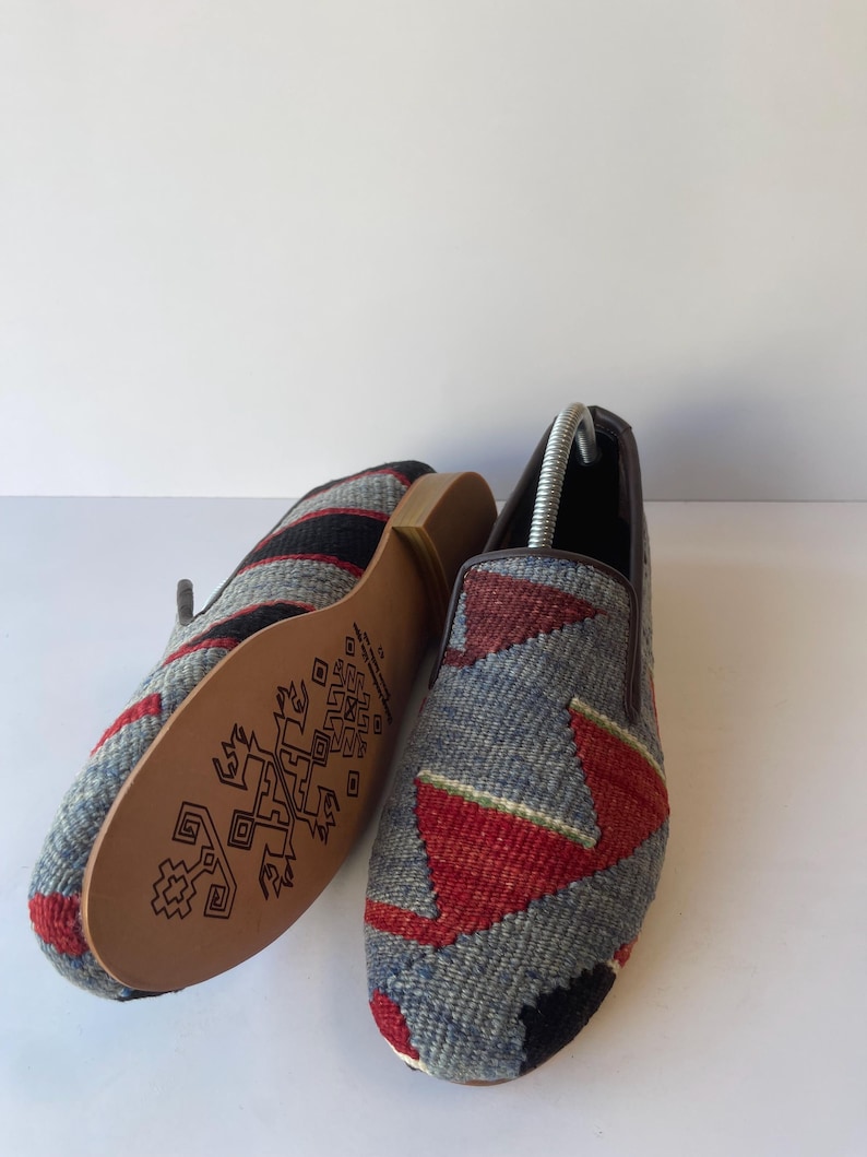 Pode incluir: Um par de sapatos slip-on tecidos com um padr&atilde;o geom&eacute;trico em cinza, vermelho e preto. As solas s&atilde;o de cor castanho claro com um design decorativo castanho escuro. Os sapatos s&atilde;o exibidos em uma superf&iacute;cie branca.