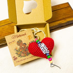 Könnte beinhalten: Ein roter gehäkelter Herz-Schlüsselanhänger mit den Worten "LOVE MAMA" in weißen Buchstabenperlen. Der Schlüsselanhänger ist an einer Pappschachtel mit herzförmigem Ausschnitt befestigt. Eine kleine Karte mit dem Wort "Matriarch" befindet sich ebenfalls in der Schachtel.