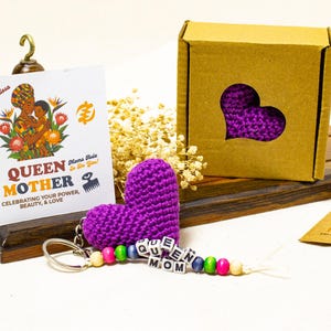 Könnte beinhalten: Ein lila gehäkelter Herz-Schlüsselanhänger mit den Worten "QUEEN MOM" auf Buchstabenperlen. Eine Karte mit dem Text "QUEEN MOTHER" und einer Illustration einer Mutter und eines Kindes wird neben einer herzförmigen Aussparungsbox angezeigt.