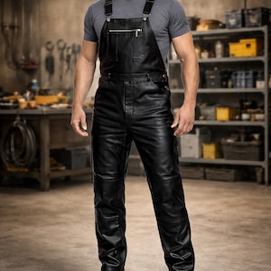 以下が含まれることがあります： 黒い革のオーバーオール。調節可能なストラップとフロントジッパーが付いています。オーバーオールは膝部分が補強されており、黒いブーツと組み合わせています。モデルはグレーのTシャツを着ています。背景はワークショップです。