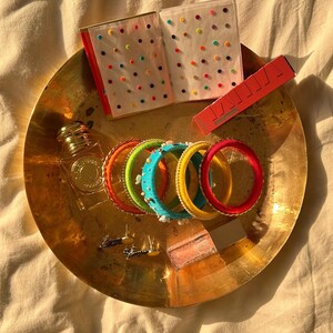 Könnte beinhalten: Eine Sammlung bunter Armreifen, Ohrringe, ein Parfümflakon und ein kleines Notizbuch mit bunten Punkten, die auf einer goldfarbenen Schale angeordnet sind. Eine rote Lippenstift-Tube ist ebenfalls vorhanden. Die Artikel sind auf einer cremefarbenen Oberfläche angeordnet.