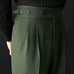 Puede incluir: Primer plano de un pantal&oacute;n verde oscuro de cintura alta con una cinturilla ancha y detalle de bot&oacute;n. El pantal&oacute;n tiene pliegues y una trabilla. El texto "TEXTHRIVE" es visible en la parte inferior de la imagen. La persona lleva un jersey negro de canal&eacute;.