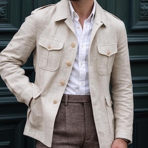 Pode incluir: Casaco de linho bege com quatro bolsos frontais e fecho de botões, usado sobre uma camisa branca abotoada e calças castanhas. O casaco tem um estilo safari clássico. O traje é adequado para um look casual elegante.