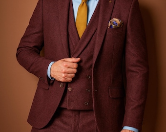 May include: A three-piece burgundy suit with a light blue shirt and mustard yellow tie. The suit includes a blazer, vest, and trousers. A patterned pocket square adds a touch of flair. The suit is made of a textured fabric.