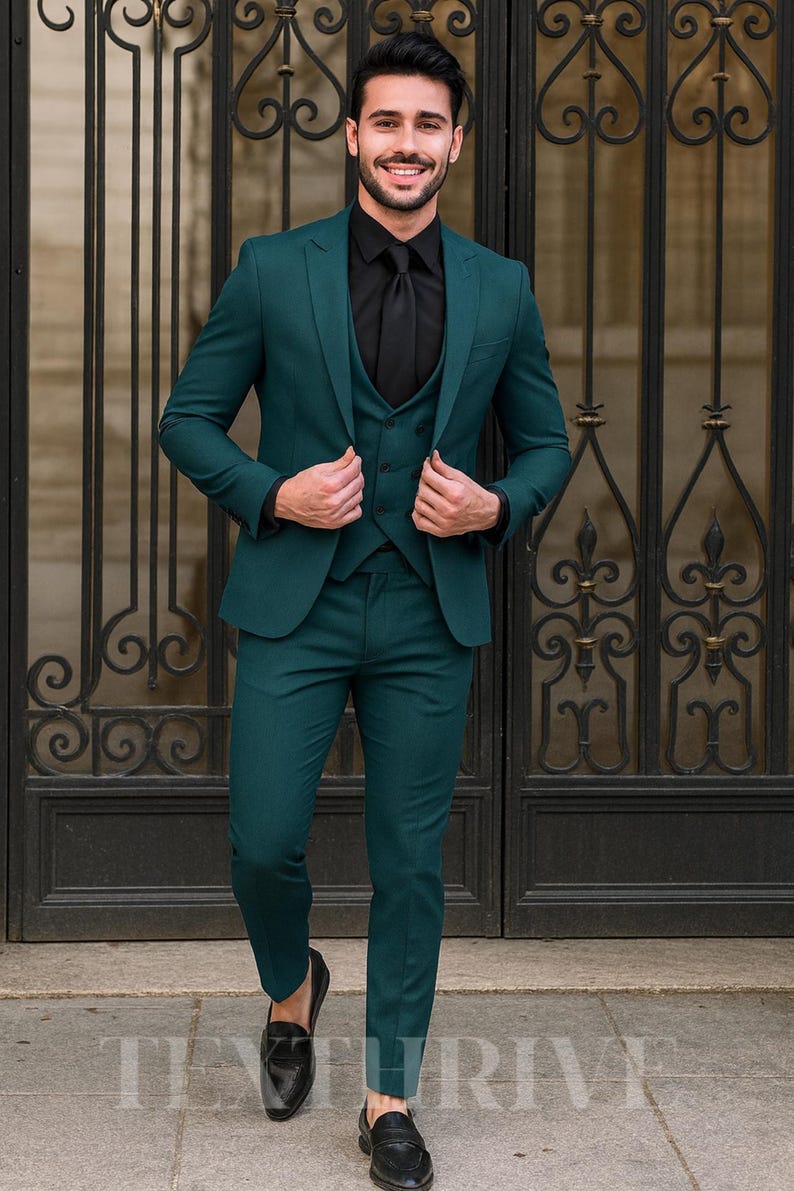 May include: A three-piece teal suit with a black shirt and tie. The suit includes a blazer, vest, and trousers. The person is wearing black loafers. The background features a decorative black metal gate.