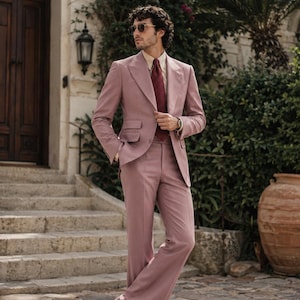 May include: A dusty rose suit featuring a single-breasted blazer with wide lapels and matching flared trousers. The ensemble is paired with a light beige button-down shirt, a dark red patterned necktie, and dark brown leather dress shoes.