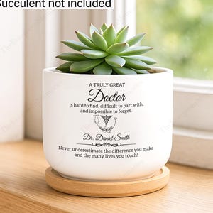 May include: White ceramic planter with a succulent, sitting on a wooden coaster. The planter has text that reads "A Truly Great Doctor" with a medical symbol and the name "Dr. Daniel Smith". The text also says "Never underestimate the difference you make and the many lives you touch!"