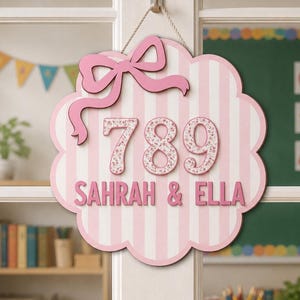 May include: A scalloped, cloud-shaped wall sign featuring vertical light pink and white stripes. The sign displays the number 789 in a floral pattern, the names SAHRAH & ELLA in pink lettering, and a pink bow accent, all suspended by a rope.