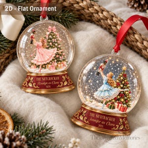 May include: Two flat, circular 2D ornaments featuring a snow globe design. One shows a girl in a pink dress holding a nutcracker, the other a girl in a blue dress. Both bases are red with gold trim and text reading The Nutcracker, names, and 2026.