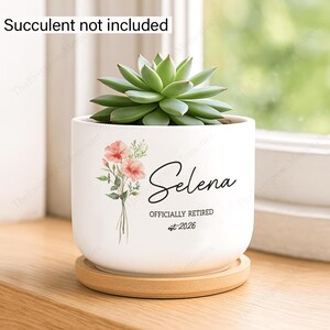 May include: White ceramic planter with a succulent, resting on a wooden saucer. The planter features a floral design and the name "Selena" with "OFFICIALLY RETIRED est. 2026". The succulent is green and the flowers are pink.