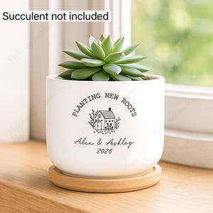 May include: A white ceramic cylindrical planter with a bamboo saucer. The front features black text reading "PLANTING NEW ROOTS", a small house illustration, the names "Alex & Ashley", and the year "2026". A text box states "Succulent not included".
