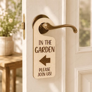 May include: A light-colored wooden door hanger with a rounded top cutout, hanging from a brass-colored door handle. The sign features dark brown engraved text that reads IN THE GARDEN, a left-pointing arrow, and the text PLEASE JOIN US!
