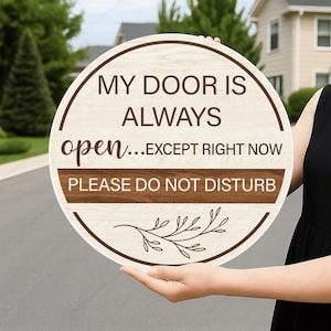 May include: A round wooden sign with a light wood grain background and a dark brown horizontal stripe. The text reads: MY DOOR IS ALWAYS open...EXCEPT RIGHT NOW PLEASE DO NOT DISTURB. A dark brown line drawing of a leafy branch is at the bottom.