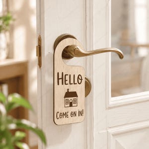 May include: A light-colored wooden door hanger with a smooth finish, featuring the dark brown text "HELLO" at the top, a simple house icon in the center, and the text "COME ON IN!" at the bottom, hanging from a brass-colored metal door handle.