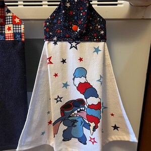 May include: A hanging kitchen towel with a navy blue top decorated with red and white stars. The white body of the towel features a cartoon character wearing sunglasses and holding a red, white, and blue ice cream cone. Red and blue stars are scattered across the towel.