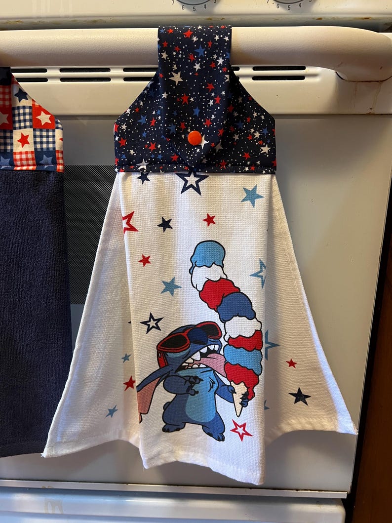 May include: A hanging kitchen towel with a navy blue top decorated with red and white stars. The white body of the towel features a cartoon character wearing sunglasses and holding a red, white, and blue ice cream cone. Red and blue stars are scattered across the towel.