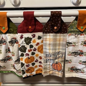 Puede incluir: Cuatro toallas de cocina con temática otoñal colgadas en un estante. Las toallas presentan diferentes diseños, que incluyen calabazas, hojas, cuadros y un camión con calabazas. Una toalla tiene el texto "Morning Pumpkin!" bordado.