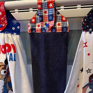 May include: Three kitchen towels hanging on an oven bar. The towels display patriotic designs in red, white, and blue. One towel has a star and plaid pattern, while the others have cartoon characters and text.