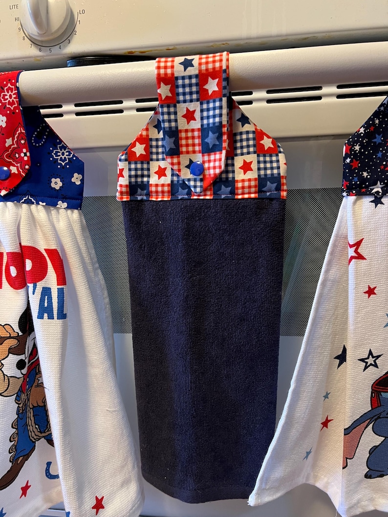 May include: Three kitchen towels hanging on an oven bar. The towels display patriotic designs in red, white, and blue. One towel has a star and plaid pattern, while the others have cartoon characters and text.